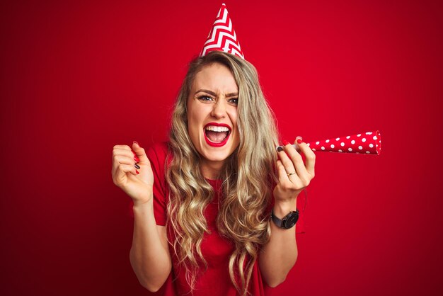 Giovane bella donna alla festa di compleanno su sfondo rosso isolato urlando orgoglioso e celebrando la vittoria e il successo commozione molto eccitata