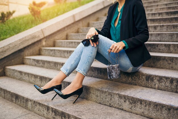 Giovane bella donna alla moda seduta in strada, indossa jeans, giacca nera, camicetta verde, occhiali da sole