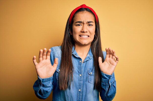 Giovane bella donna asiatica che indossa camicia di jeans casual e diadema su sfondo giallo espressione disgustata dispiaciuta e timorosa facendo faccia disgustata perché reazione di avversione con le mani alzate