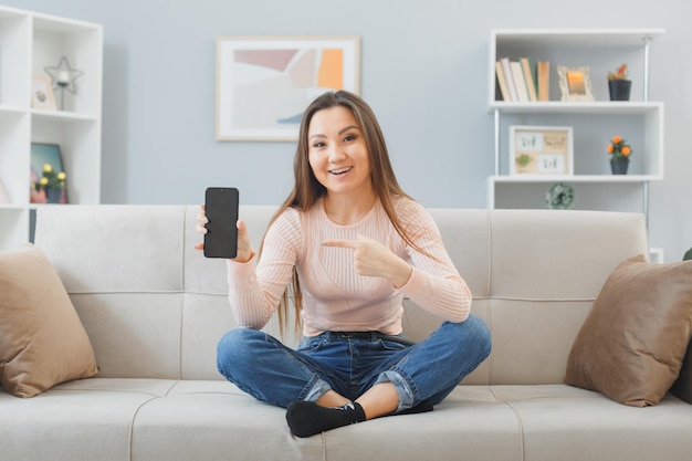 Giovane bella donna asiatica in abiti da casa seduta su un divano a casa interno che mostra smartphone che punta con il dito indice su di esso guardando sorridente sicuro di trascorrere del tempo a casa