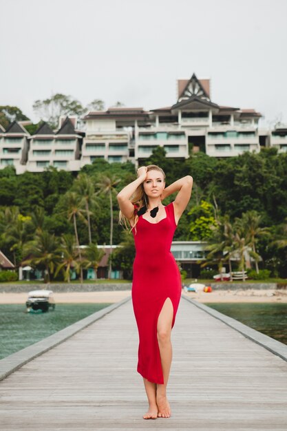Giovane bella donna attraente in piedi da solo sul molo in hotel resort di lusso, vacanze estive, abito lungo rosso, capelli biondi, abbigliamento sexy, spiaggia tropicale, seducente, sensuale, sorridente