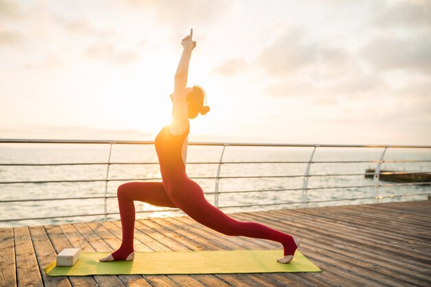 Giovane bella donna attraente sottile che fa yoga al mattino all'alba dal mare, stile di vita sano