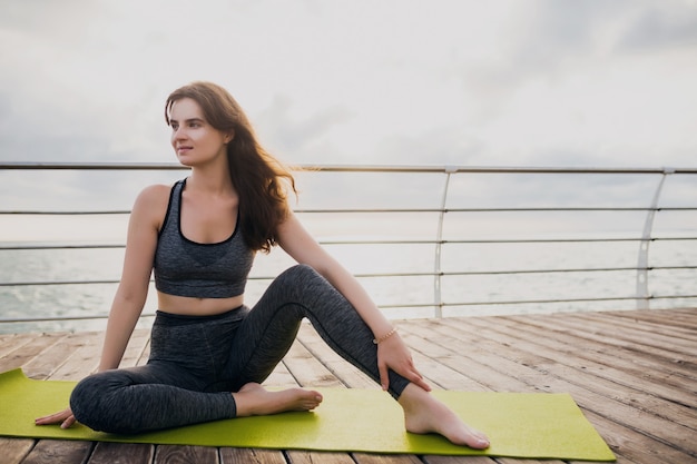 Giovane bella donna attraente sottile che si distende sulla stuoia di yoga al mattino all'alba dal mare, stile di vita sano, sport fitness