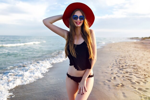 Giovane bella donna bionda che indossa un bikini nero, corpo snello, godersi le vacanze e divertirsi sulla spiaggia, lunghi capelli biondi, occhiali da sole e cappello di paglia. Vacanze a Bali.