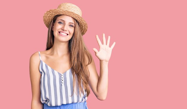 Giovane bella donna bionda che indossa un cappello estivo che mostra e punta verso l'alto con le dita numero cinque mentre sorride fiducioso e felice.
