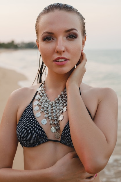 Giovane bella donna bionda che prende il sole sulla spiaggia di sabbia in costume da bagno bikini, collana vintage