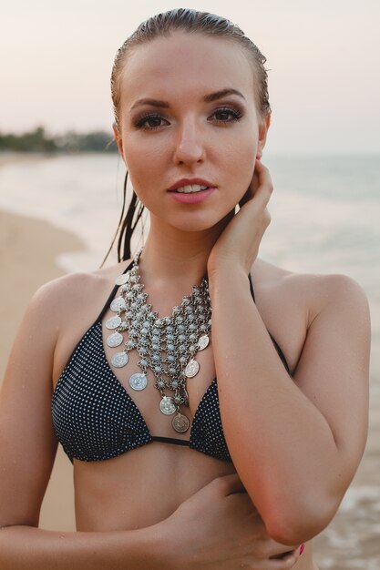 Giovane bella donna bionda che prende il sole sulla spiaggia di sabbia in costume da bagno bikini, collana vintage