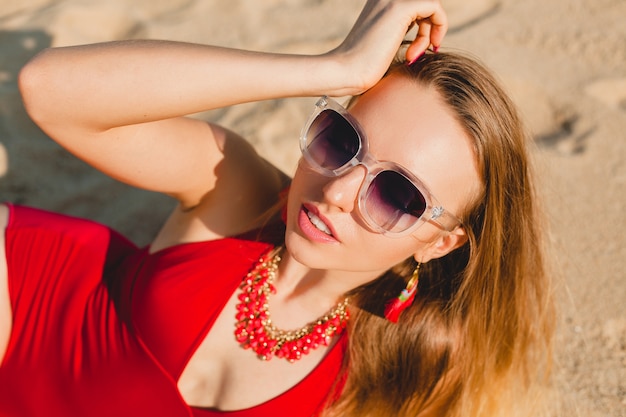 Giovane bella donna bionda che prende il sole sulla spiaggia di sabbia in costume da bagno rosso, occhiali da sole