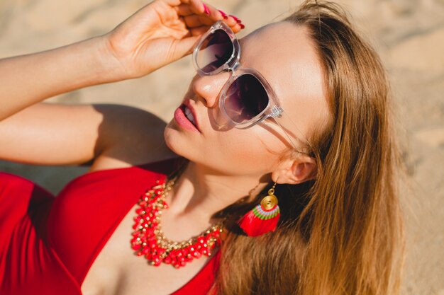 Giovane bella donna bionda che prende il sole sulla spiaggia di sabbia in costume da bagno rosso, occhiali da sole