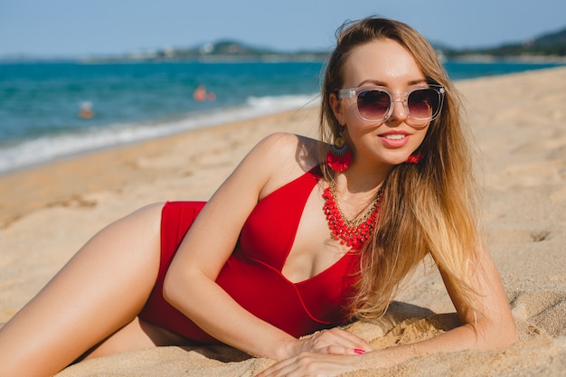 Giovane bella donna bionda che prende il sole sulla spiaggia di sabbia in costume da bagno rosso, occhiali da sole
