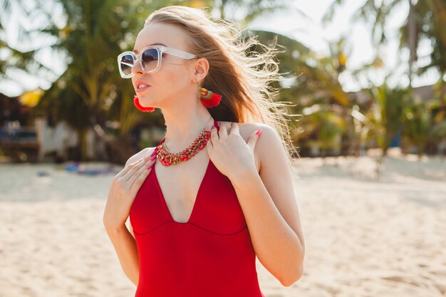 Giovane bella donna bionda che prende il sole sulla spiaggia in costume da bagno rosso, occhiali da sole