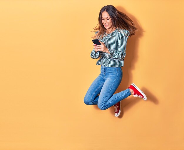 Giovane bella donna bionda che salta con il sorriso sul viso. Utilizzo dello smartphone su sfondo giallo isolato