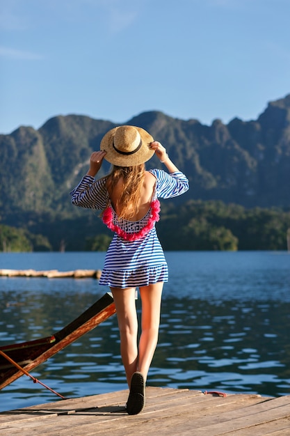 Giovane bella donna bionda con i capelli lunghi, che indossa un cappello vintage e vestito alla moda sexy luminoso guarda alle montagne e al lago, mostra la sua mano, incredibili avventure estive.