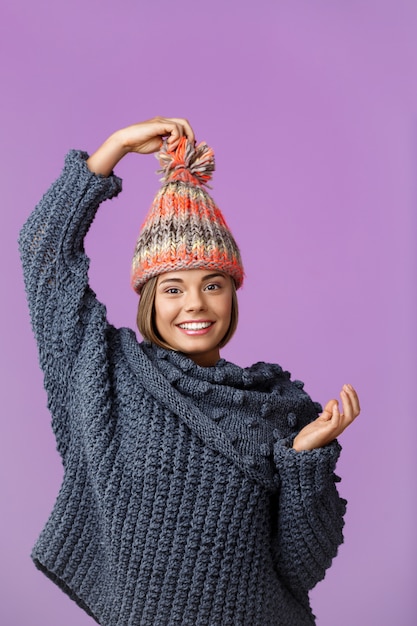 Giovane bella donna bionda divertente in cappello lavorato a maglia e maglione sorridente in posa sulla viola. Copia spazio.
