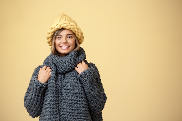 Giovane bella donna bionda in cappello e maglione tricottati che sorride sul giallo.