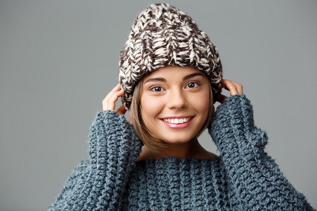 Giovane bella donna bionda in cappello e maglione tricottati che sorride sul gray.