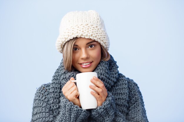 Giovane bella donna bionda in cappello e maglione tricottati che tengono tazza che sorride sul blu.