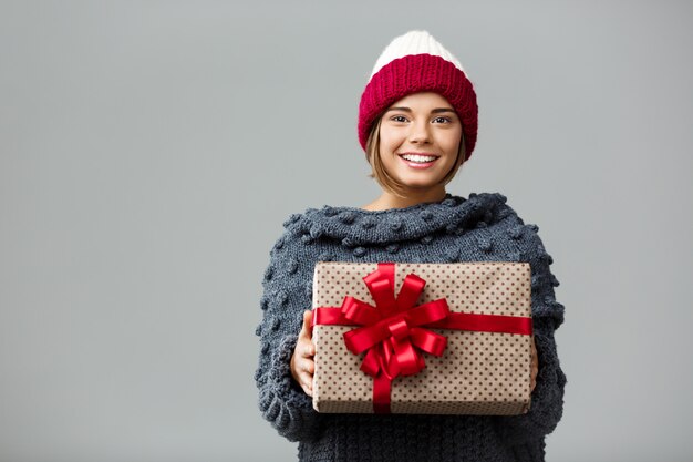 Giovane bella donna bionda in cappello e maglione tricottati che tiene il contenitore di regalo sorridente su gray.