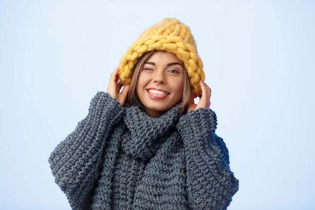 Giovane bella donna bionda in cappello lavorato a maglia e maglione che sbatte le palpebre sorridente sul blu.