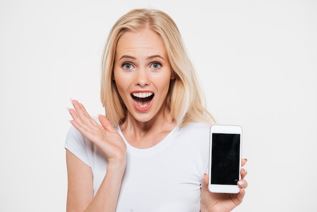 Giovane bella donna bionda sorpresa con la bocca aperta, mostrando lo schermo in bianco dello smartphone