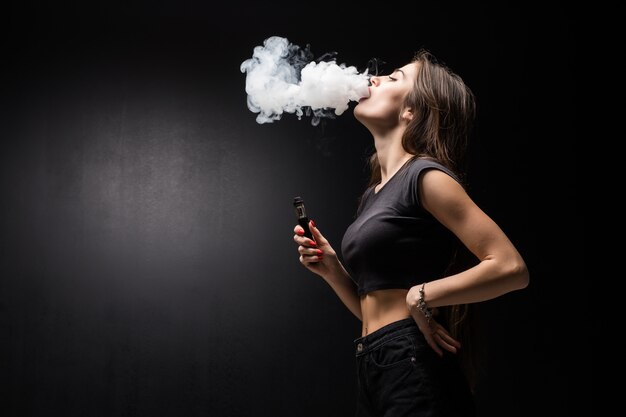 Giovane bella donna castana che fuma, vaping sigaretta elettronica con fumo sulla parete nera