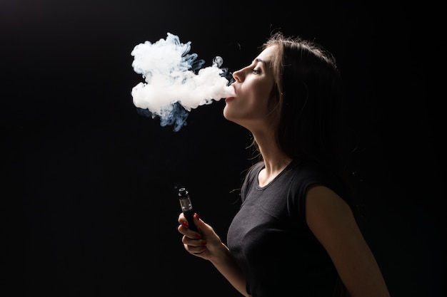 Giovane bella donna castana che fuma, vaping sigaretta elettronica con fumo sulla parete nera