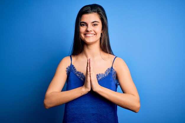 Giovane bella donna castana che indossa biancheria intima blu elegante e confortevole che prega con le mani insieme chiedendo perdono sorridendo fiducioso