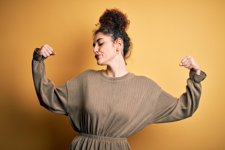 Foto gratuito giovane bella donna castana con capelli ricci e piercing che indossa un abito casual che mostra i muscoli delle braccia sorridendo orgoglioso concetto di fitness