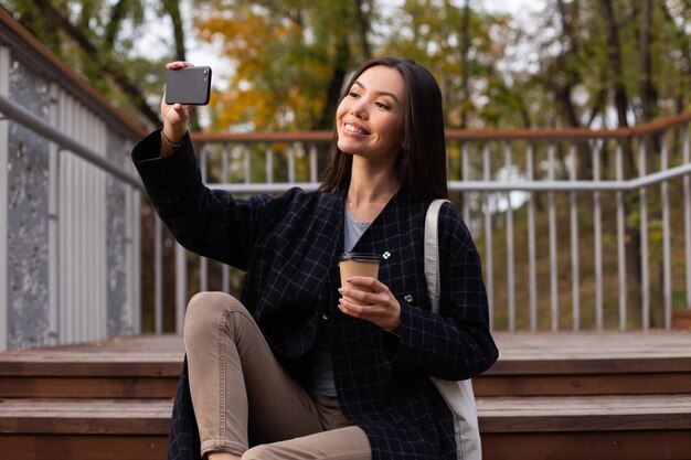 Giovane bella donna casual con caffè per andare felicemente prendendo selfie sul cellulare nel parco autunnale