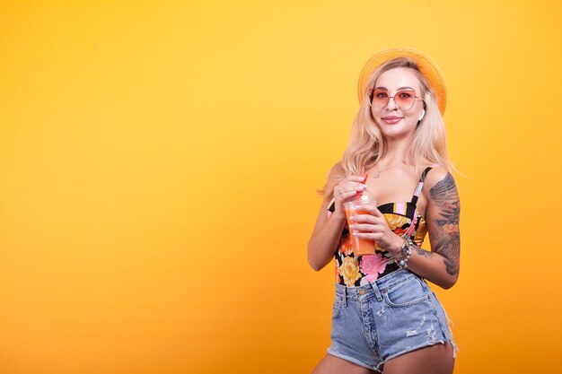 Giovane bella donna che beve succo d'arancia fresco con cappello e occhiali da sole in studio