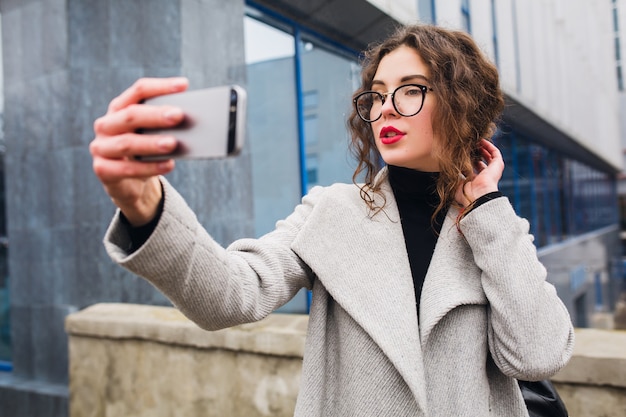 Giovane bella donna che cammina per la strada della città in cappotto grigio, stile moda autunno, occhiali, tenendo lo smartphone, scattare foto selfie, sorridente, felice