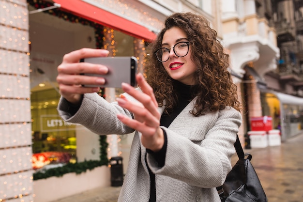 Giovane bella donna che cattura foto seflie utilizzando smartphone, autunno street city style, cappotto caldo, occhiali, felice, sorridente, tenendo il telefono in mano, capelli ricci