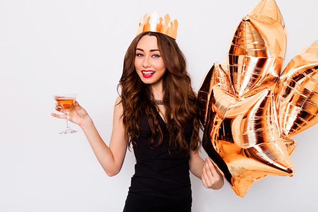 Giovane bella donna che celebra in abito nero sorriso e in posa con cocktail in mano e palloncini di purezza.