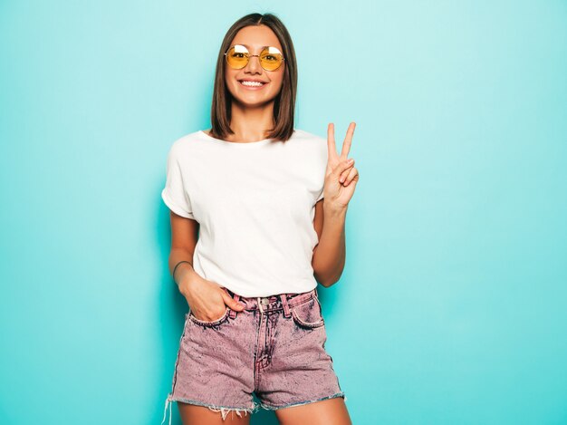 Giovane bella donna che guarda l'obbiettivo. Ragazza alla moda in pantaloncini di maglietta e jeans bianchi estivi casual in occhiali da sole rotondi. La femmina positiva mostra le emozioni facciali. Il modello isolato sul blu mostra il segno di pace