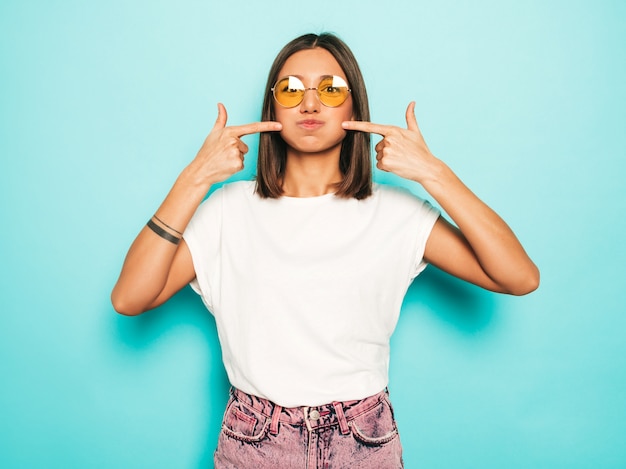 Giovane bella donna che guarda l'obbiettivo. Ragazza alla moda in pantaloncini di maglietta e jeans bianchi estivi casual in occhiali da sole rotondi. La femmina positiva mostra le emozioni facciali. Modello divertente che soffia le sue guance.