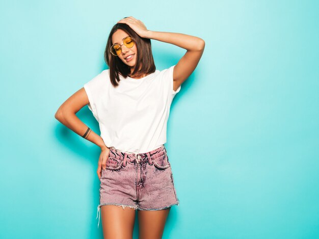 Giovane bella donna che guarda l'obbiettivo. Ragazza alla moda in pantaloncini di maglietta e jeans bianchi estivi casual in occhiali da sole rotondi. La femmina positiva mostra le emozioni facciali. Modello divertente isolato sul blu