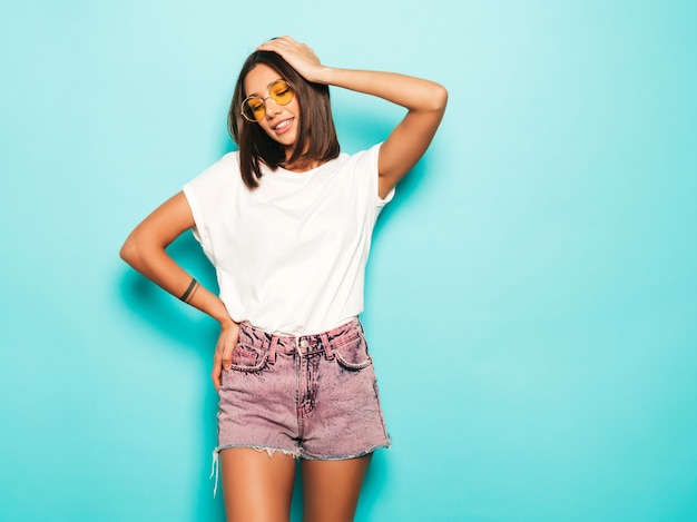 Giovane bella donna che guarda l'obbiettivo. Ragazza alla moda in pantaloncini di maglietta e jeans bianchi estivi casual in occhiali da sole rotondi. La femmina positiva mostra le emozioni facciali. Modello divertente isolato sul blu