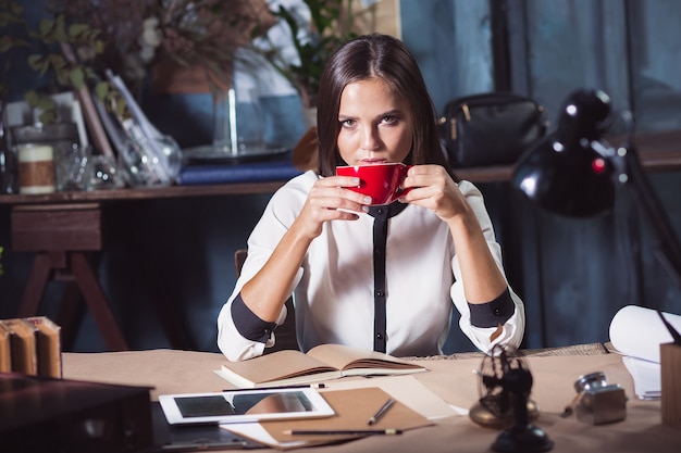 Giovane bella donna che lavora con una tazza di caffè e un taccuino in ufficio loft