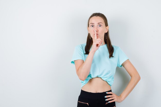 Giovane bella donna che mostra gesto di silenzio in maglietta, pantaloni e sguardo concentrato. vista frontale.