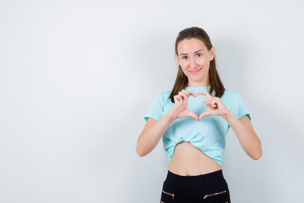 Giovane bella donna che mostra il gesto del cuore in maglietta e sembra allegra, vista frontale.