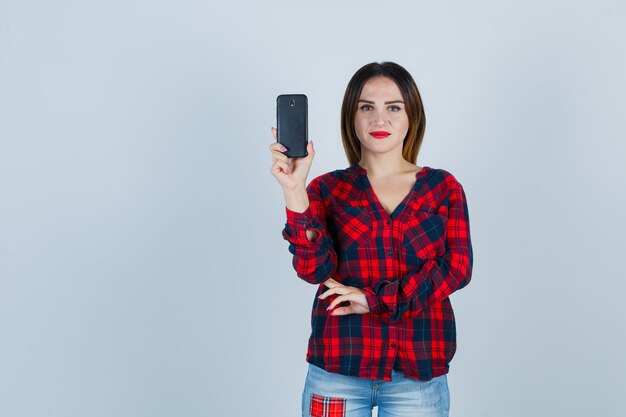 Giovane bella donna che mostra telefono in camicia casual e sembra allegra. vista frontale.