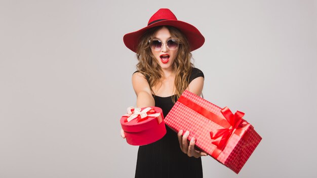Giovane bella donna che tiene i regali, vestito nero, cappello rosso, occhiali da sole, felice, sorridente, sexy, elegante, scatole regalo, celebrando, positivo, emotivo