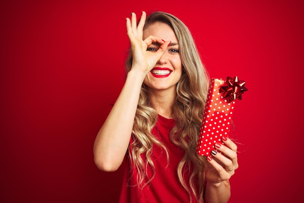 Giovane bella donna che tiene il regalo di San Valentino su sfondo rosso isolato con faccia felice sorridente facendo segno ok con la mano sull'occhio guardando attraverso le dita