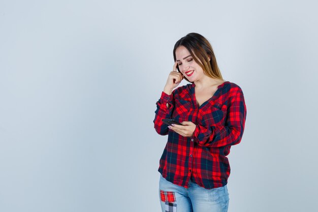 Giovane bella donna che tiene il telefono, tenendo la mano sul viso in camicia casual e sembra allegra, vista frontale.