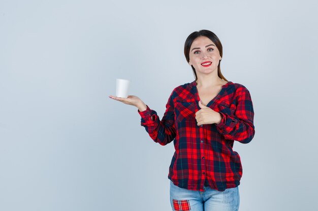 Giovane bella donna che tiene in mano un bicchiere di plastica, che mostra pollice in su in camicia casual, jeans e sembra allegra, vista frontale.