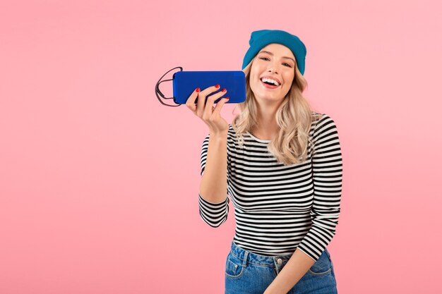 Giovane bella donna con altoparlante wireless che ascolta musica indossando camicia a righe e cappello blu sorridente felice umore positivo in posa su sfondo rosa