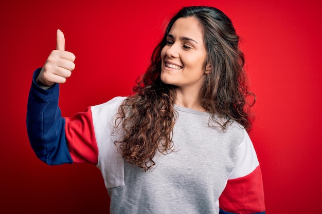 Giovane bella donna con capelli ricci che indossa una felpa casual su sfondo rosso isolato Guardando fiero sorridente facendo pollice in alto gesto a lato