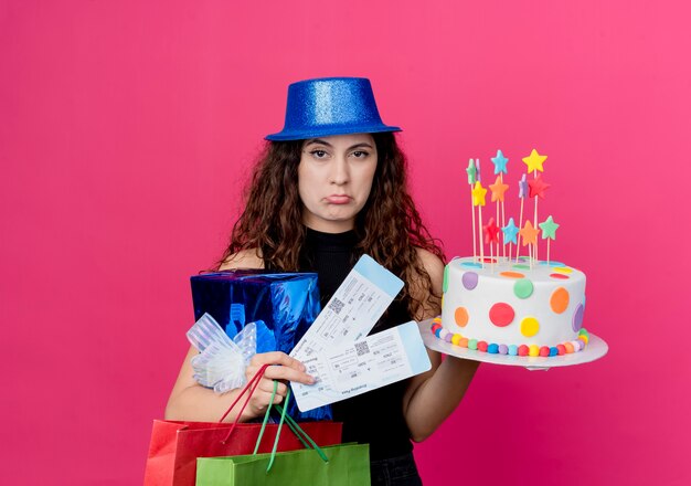 Giovane bella donna con capelli ricci in un cappello di festa che tiene il contenitore di regalo della torta di compleanno e biglietti aerei con il concetto di festa di compleanno di espressione triste in piedi sopra il muro rosa