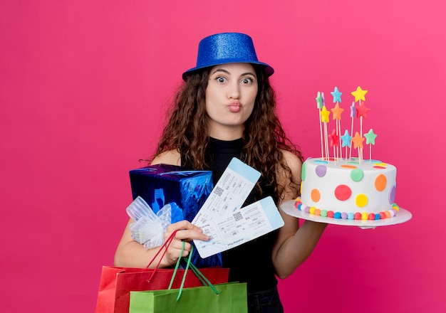 Giovane bella donna con capelli ricci in un cappello di festa che tiene il contenitore di regalo della torta di compleanno e biglietti aerei concetto di festa di compleanno felice e positivo in piedi sopra il muro rosa