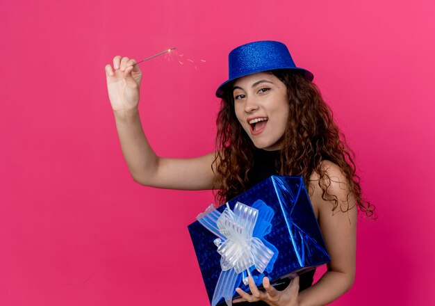 Giovane bella donna con capelli ricci in un cappello di festa che tiene il contenitore di regalo di compleanno e il concetto felice ed emozionante della festa di compleanno dello sparkler sopra il rosa